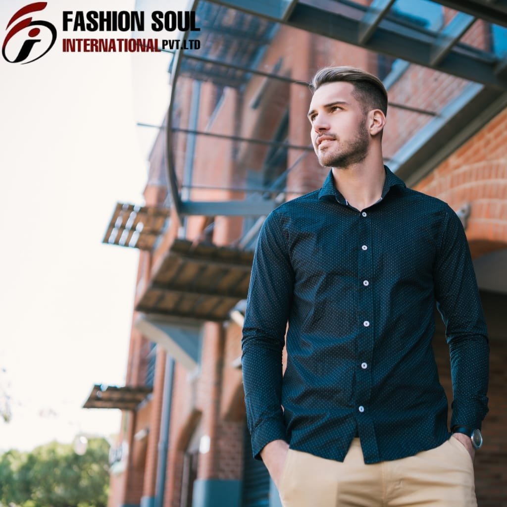  A young man with neatly styled hair and a trimmed beard is standing outdoors next to a brick building with metal fire escape stairs. He is dressed in a dark blue, long-sleeved, button-down shirt and beige pants, with his hands in his pockets, gazing thoughtfully into the distance.