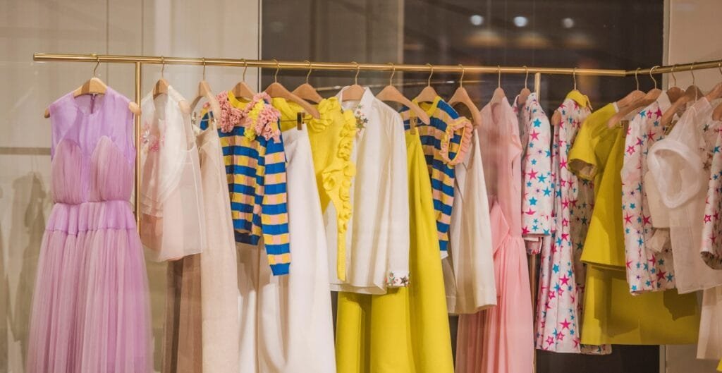 A row of colorful dresses and children's clothing hanging on wooden hangers on a gold clothing rack inside a store.