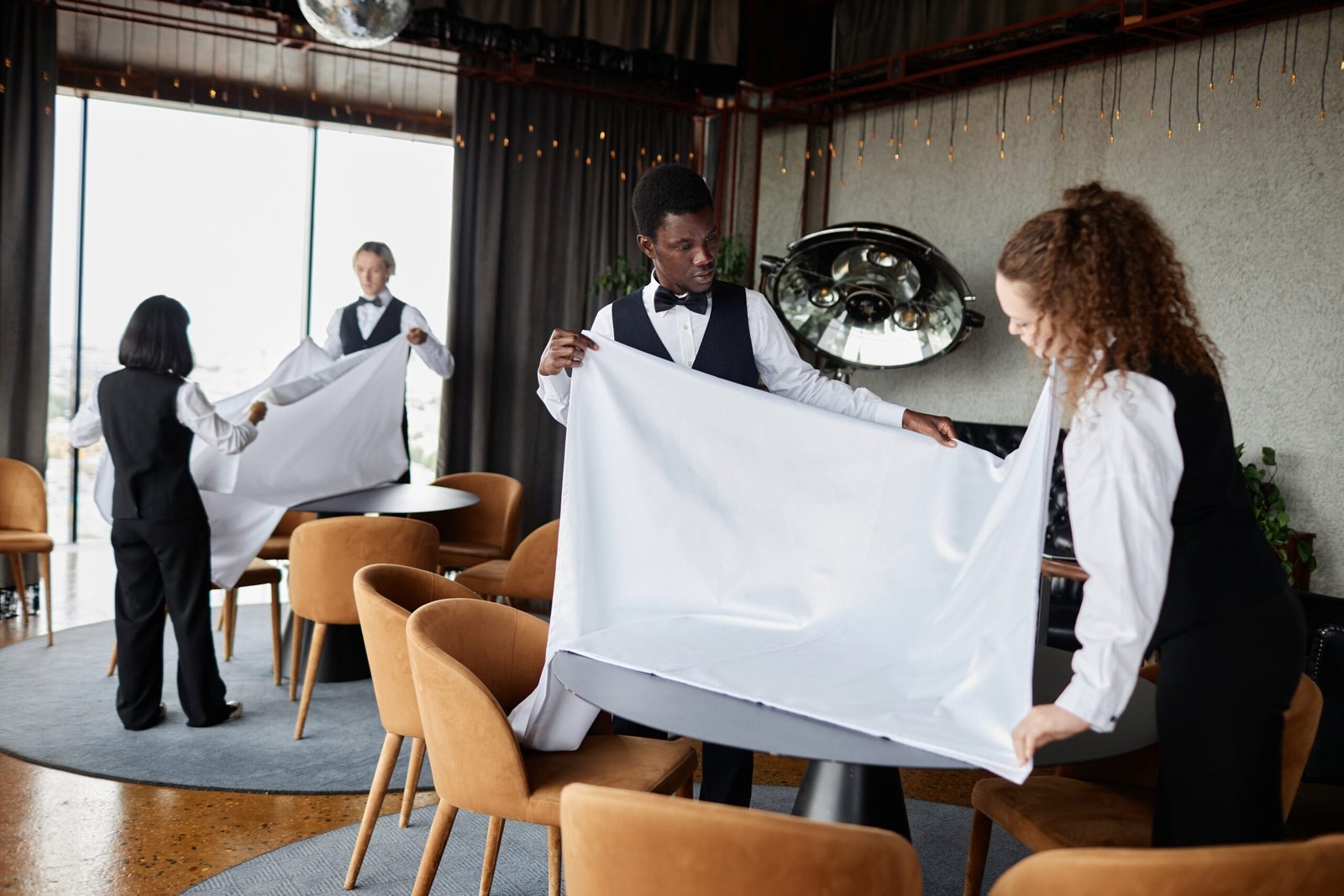 Four waitstaff folding white tablecloths in a stylish restaurant or event space with orange chairs, large window, and string lights.
