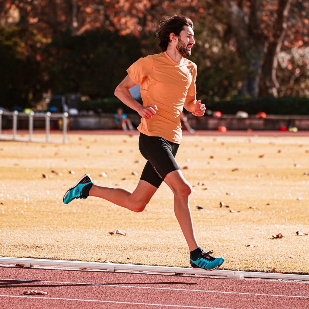 A male runner in a bright orange T-shirt and black shorts, running on an outdoor track with autumn trees in the background.