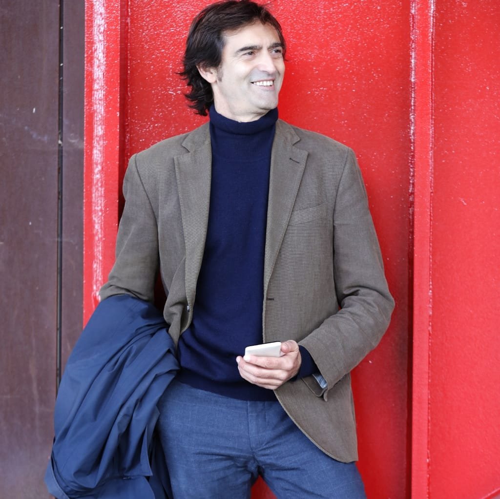 A man wearing a navy blue turtleneck sweater paired with a brown sport coat, holding a jacket in his right hand, standing against a red wall and smiling.