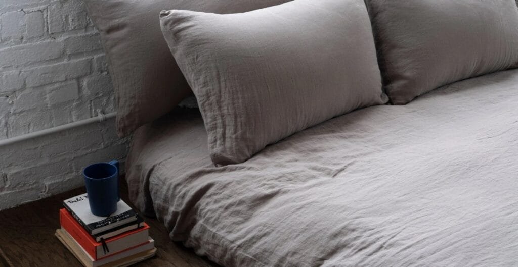 A neatly made bed with light gray bedding and pillows, positioned next to a small side table holding a stack of books and a blue cup, against a white brick wall.
