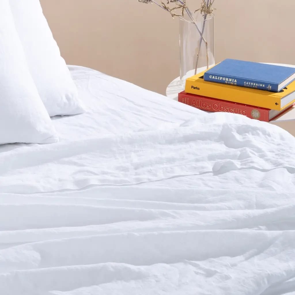 A neatly made bed with white bedding and pillows, a small round side table holding colorful books and a vase with dried flowers, set against a plain wall.