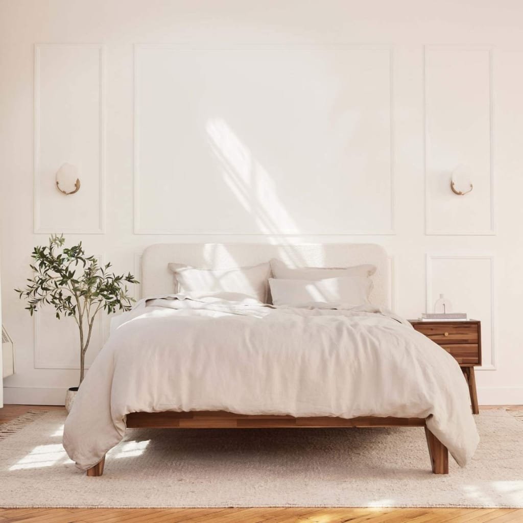 A minimalist bedroom with a large bed dressed in white linens, a wooden nightstand on the right, a potted plant on the left, and soft natural light streaming through a window, casting shadows on the wall.