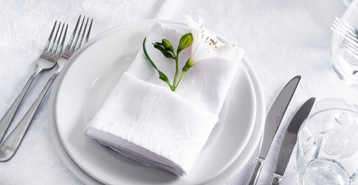 A neatly arranged table setting with a white plate holding a napkin folded with a small white flower and green leaves, surrounded by silverware and clear glasses on a white tablecloth.