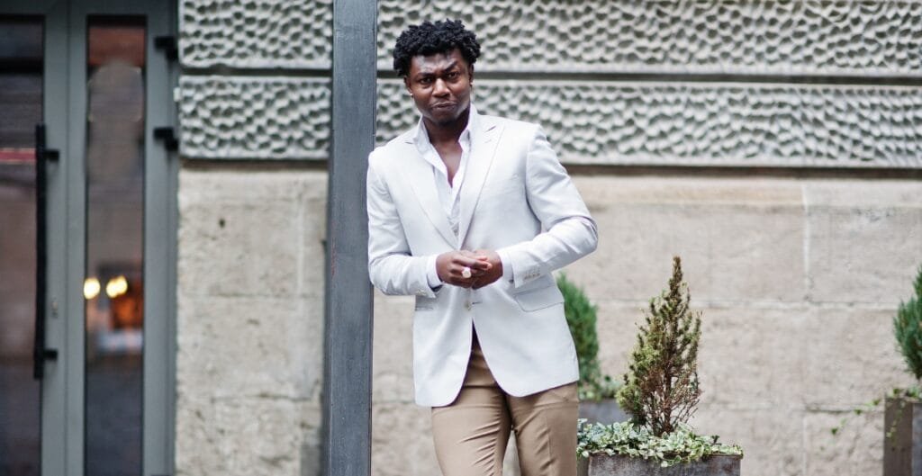 A man with curly black hair wearing a white blazer and beige pants stands outdoors, leaning against a pole with a serious expression.