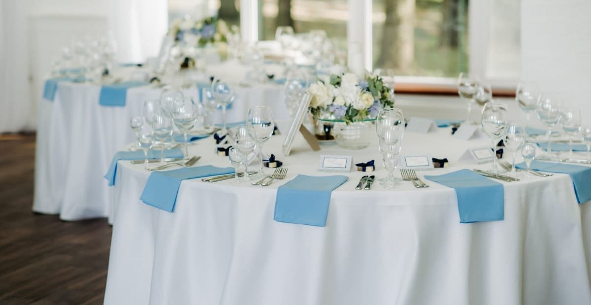 Elegant round banquet table set for a formal event, decorated with white tablecloths, light blue napkins, multiple wine glasses, and a floral centerpiece, with natural light coming through the windows.
