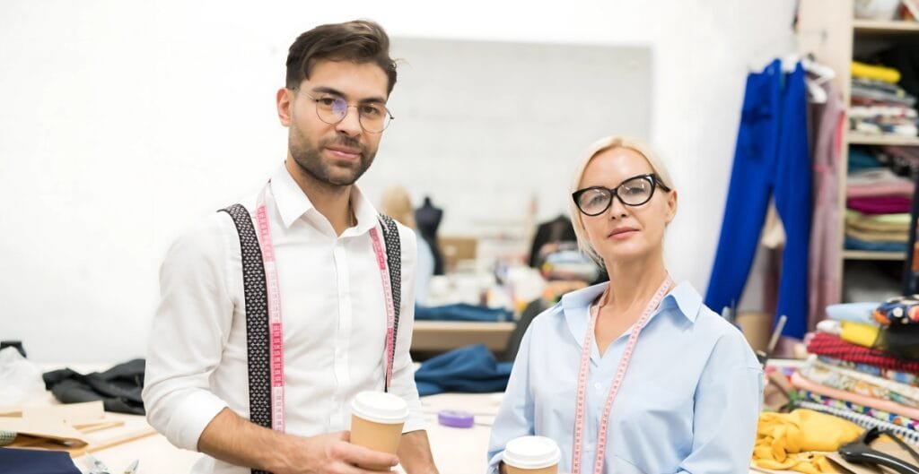 Two fashion designers or tailors standing in a clothing workshop, holding coffee cups, with measuring tapes around their necks. The background shows shelves filled with fabrics and sewing supplies.