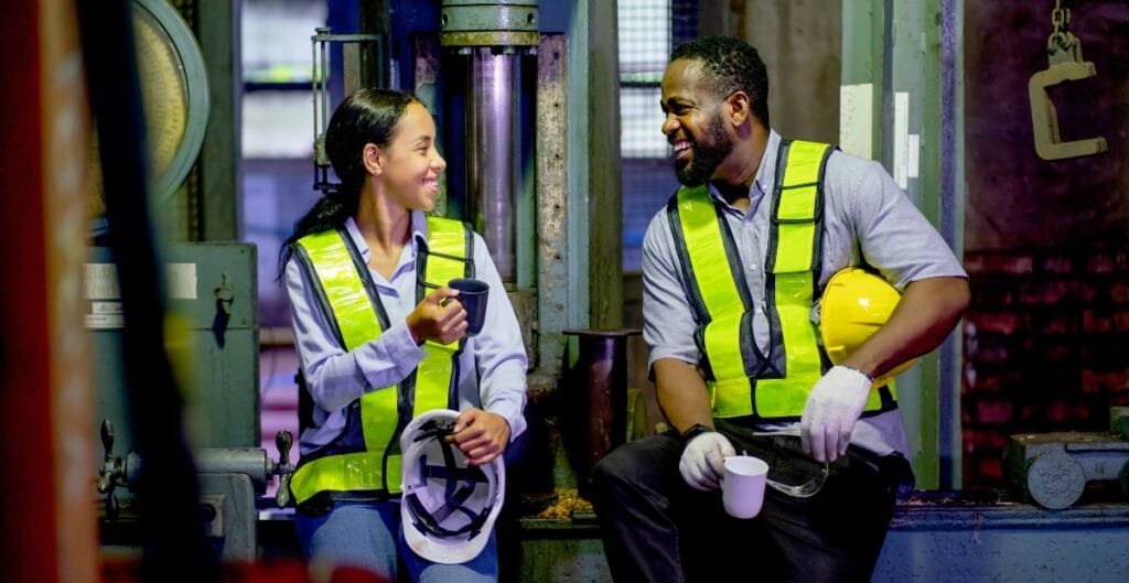 Two workers in safety vests and gloves are smiling and chatting in an industrial setting, with one holding a yellow helmet and the other holding a mug.