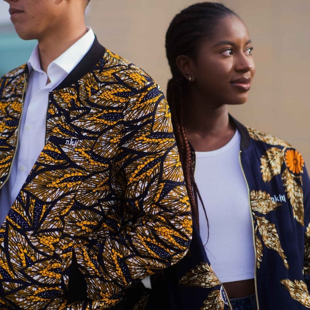 Two people standing back-to-back, wearing matching patterned jackets with yellow, navy, and white designs, with one person facing left and the other facing right, against a neutral background.