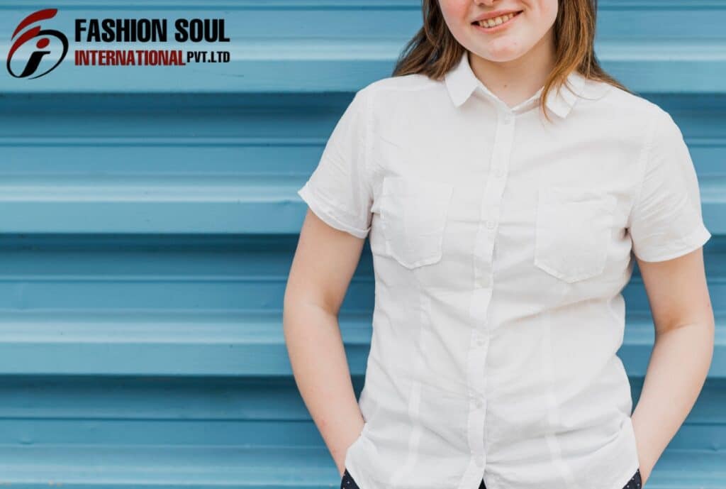 A woman with light brown hair, wearing a white short-sleeve button-up shirt, stands in front of a blue, horizontally-striped background with her hands in her pockets, smiling slightly.
