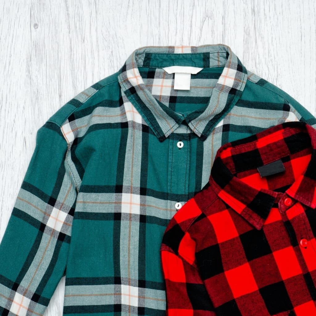 Two plaid flannel shirts on a wooden surface: a teal and gray plaid shirt layered with a red and black plaid shirt.