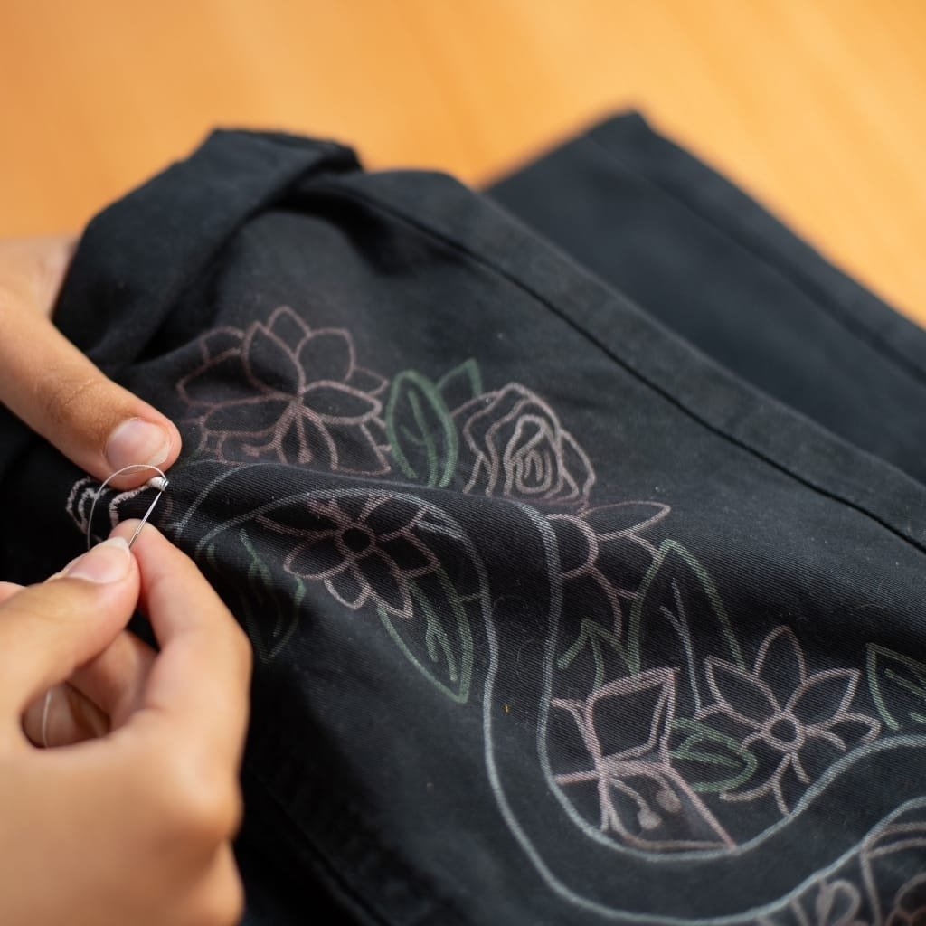 Close-up of a person sewing or embroidering a design onto a black fabric with colorful floral patterns. The person's hands are holding a needle and thread, carefully working on the fabric.