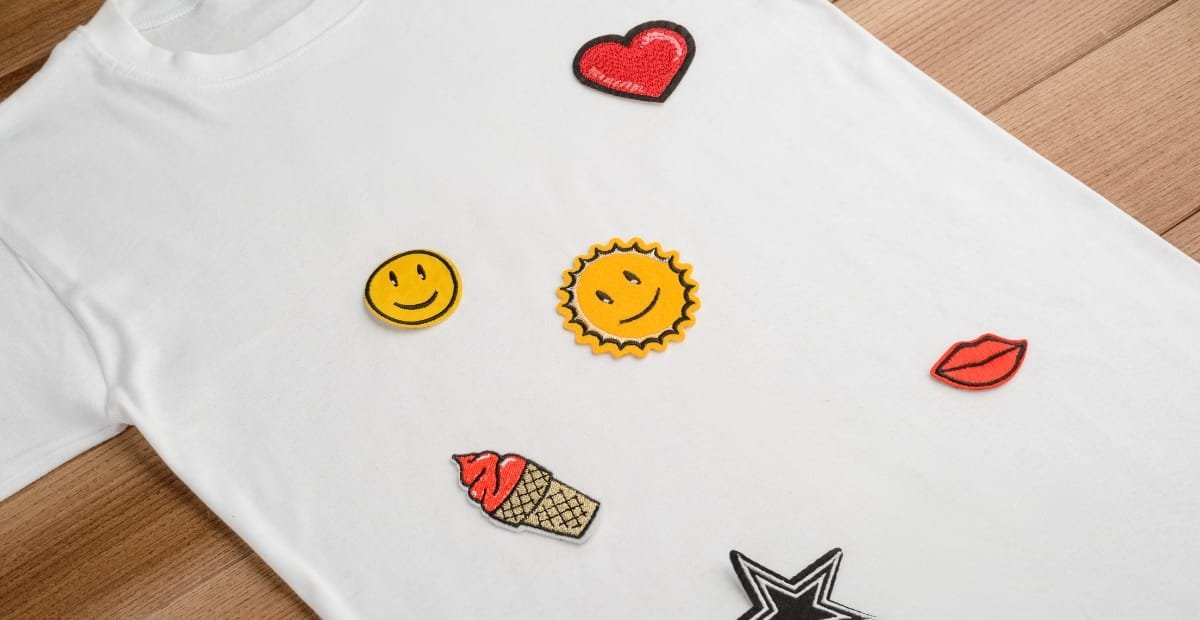 A white T-shirt laid flat on a wooden surface with various colorful embroidered patches scattered on it, including a red heart, a yellow smiley face, a sun, red lips, an ice cream cone, and a black-and-white star.