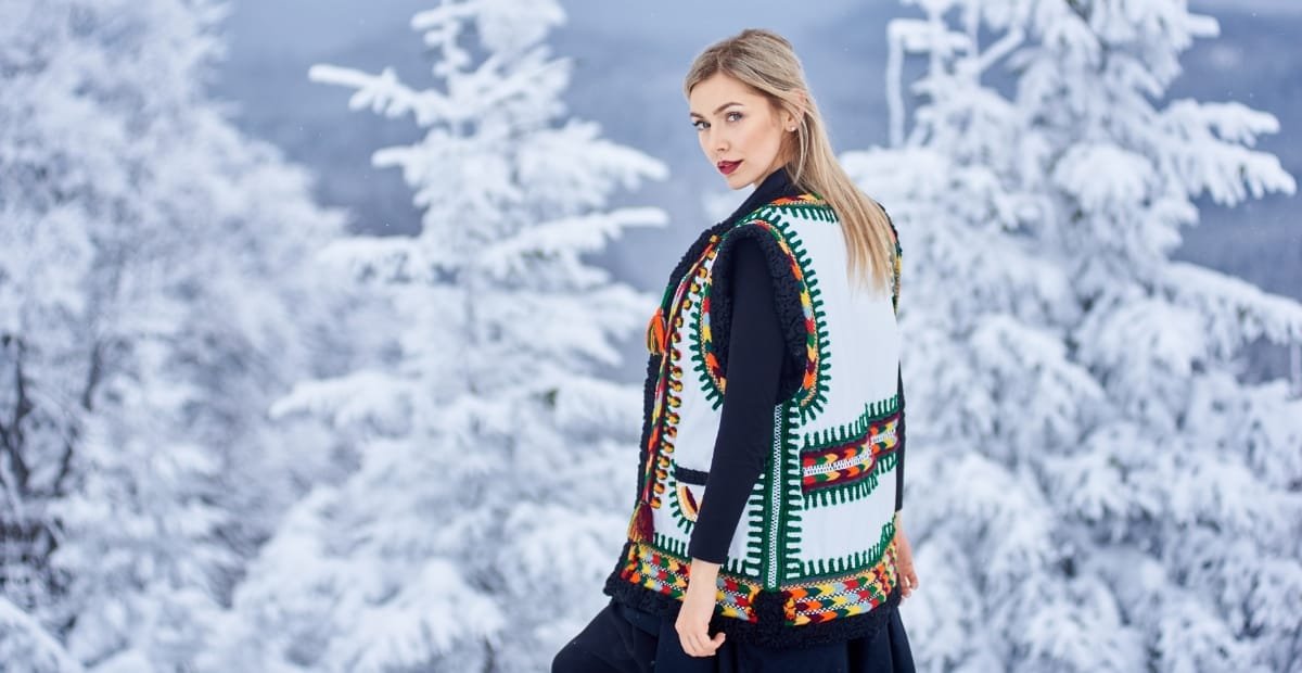 A young woman with blonde hair wearing a colorful, embroidered vest over a black outfit, standing outdoors in a snowy winter landscape with snow-covered trees in the background.