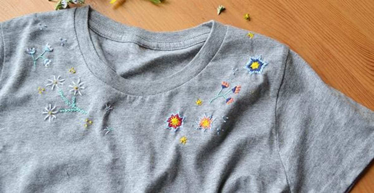 A gray T-shirt with colorful embroidered floral and star designs near the shoulder and upper chest area, placed on a wooden surface.