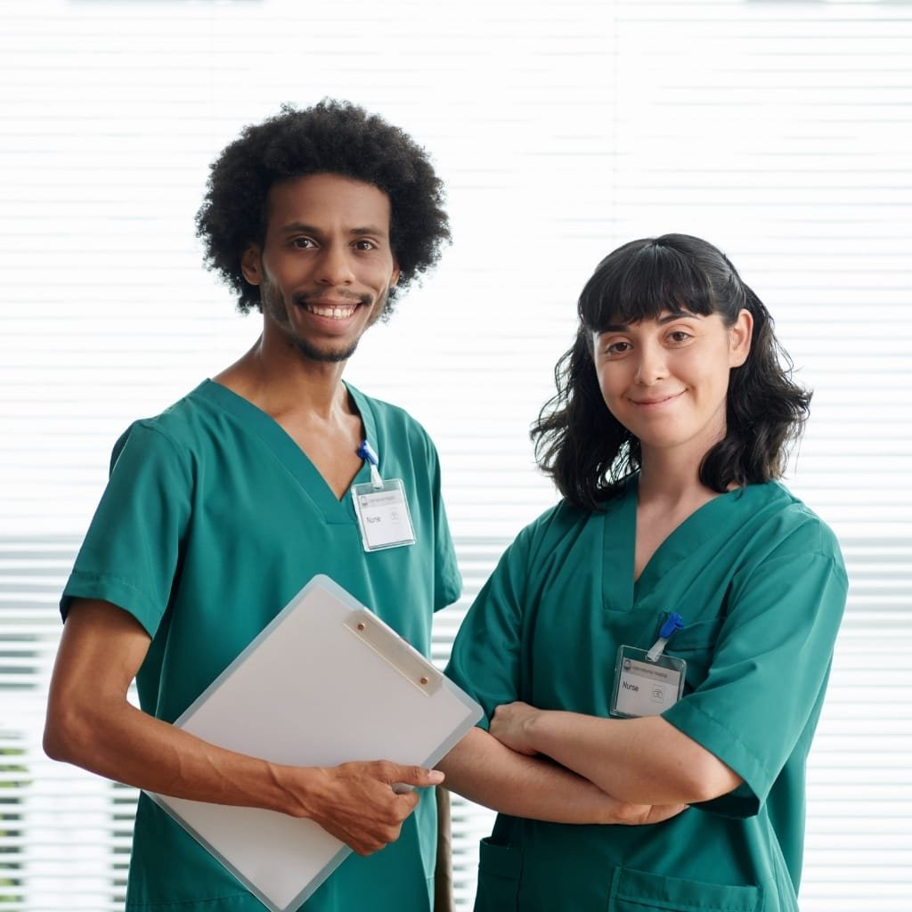Two healthcare professionals, a man and a woman, dressed in green scrubs, standing side by side with the man holding a clipboard and both smiling confidently in front of a window with blinds.