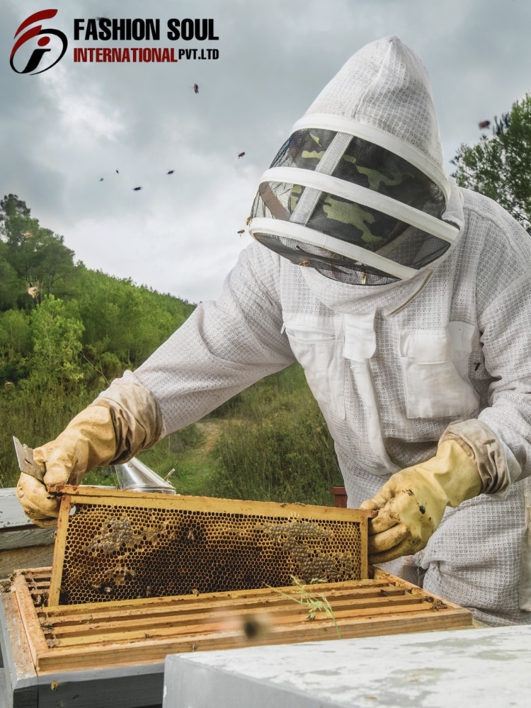 A beekeeper in full protective suit with a veil, gloves, and a camo-pattern inside the mesh, tending a wooden beehive frame outdoors. Hills and trees are in the background under a cloudy sky, with bees flying nearby.