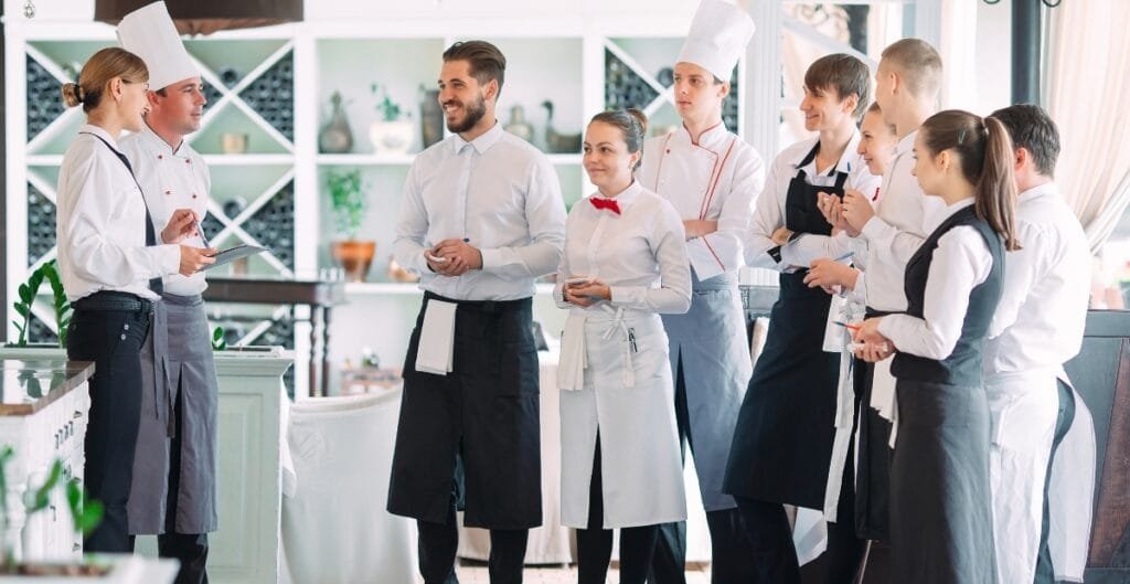 A group of restaurant staff and chefs stand in a bright dining area, wearing uniforms with aprons. They appear to be listening to a team briefing, with one man in a white shirt speaking and others smiling and holding tablets or menus.