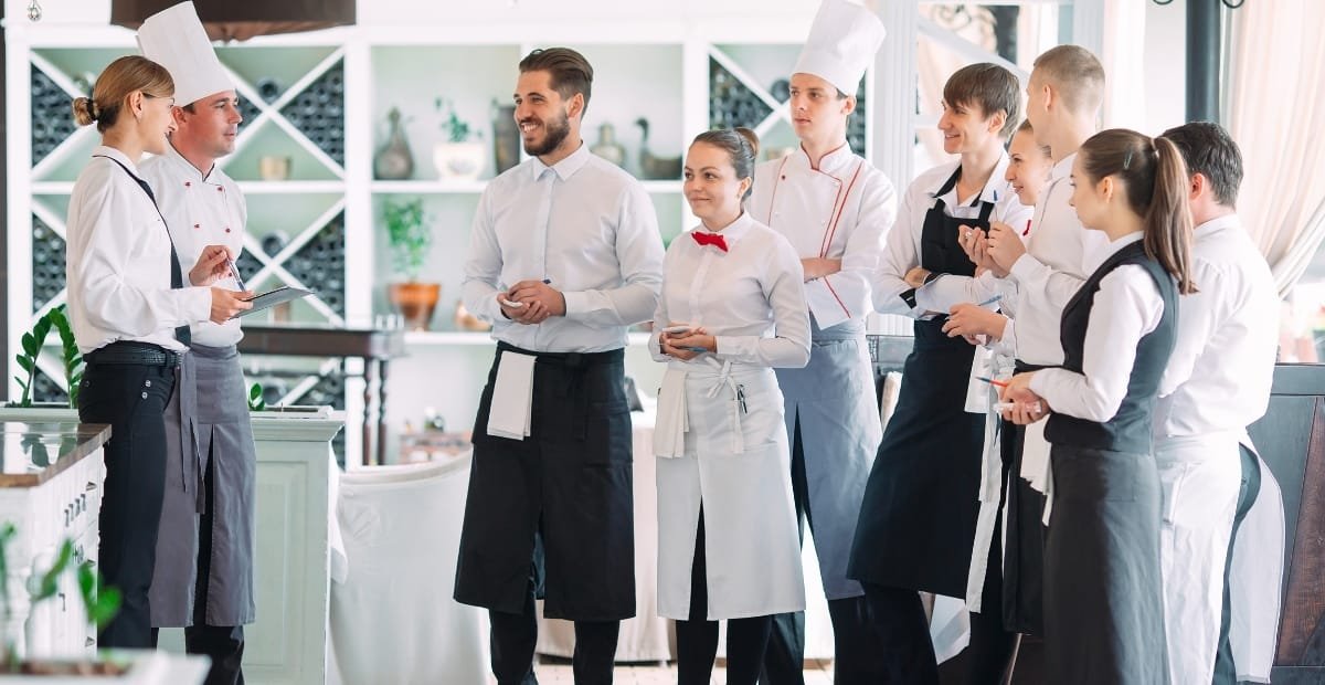 A group of restaurant staff and chefs stand in a bright dining area, wearing uniforms with aprons. They appear to be listening to a team briefing, with one man in a white shirt speaking and others smiling and holding tablets or menus.