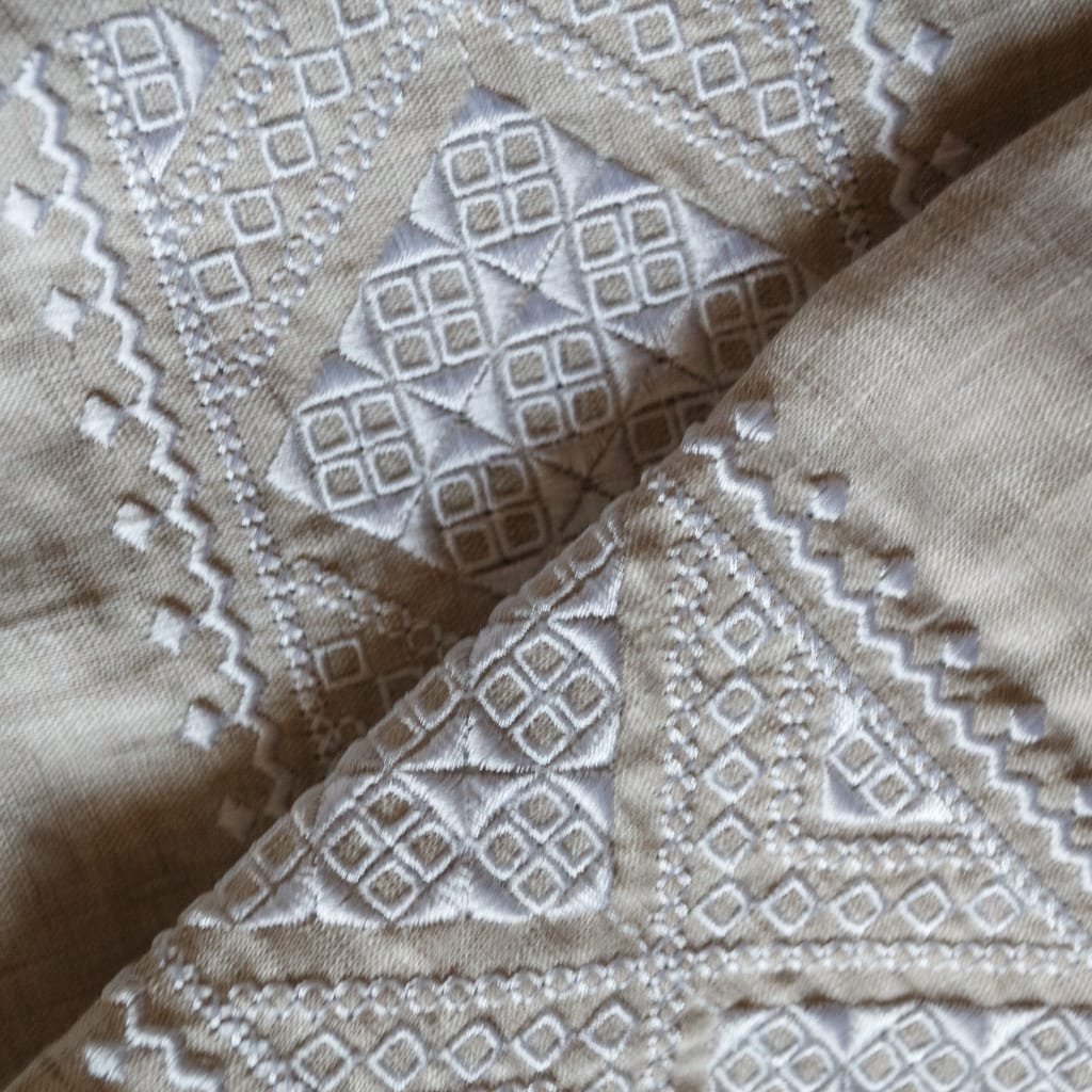 Close-up of beige fabric with intricate white embroidery forming geometric diamond and square patterns along triangular panels.