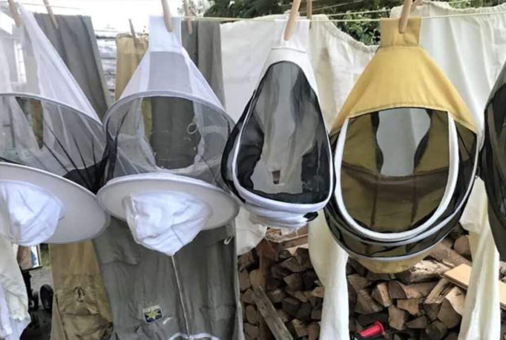 A row of mosquito netting hoods hanging on a clothesline to dry. Each hood has a mesh face opening and a solid-colored fabric top, with white rims and zippers. In the background there are more fabrics and a stack of firewood.