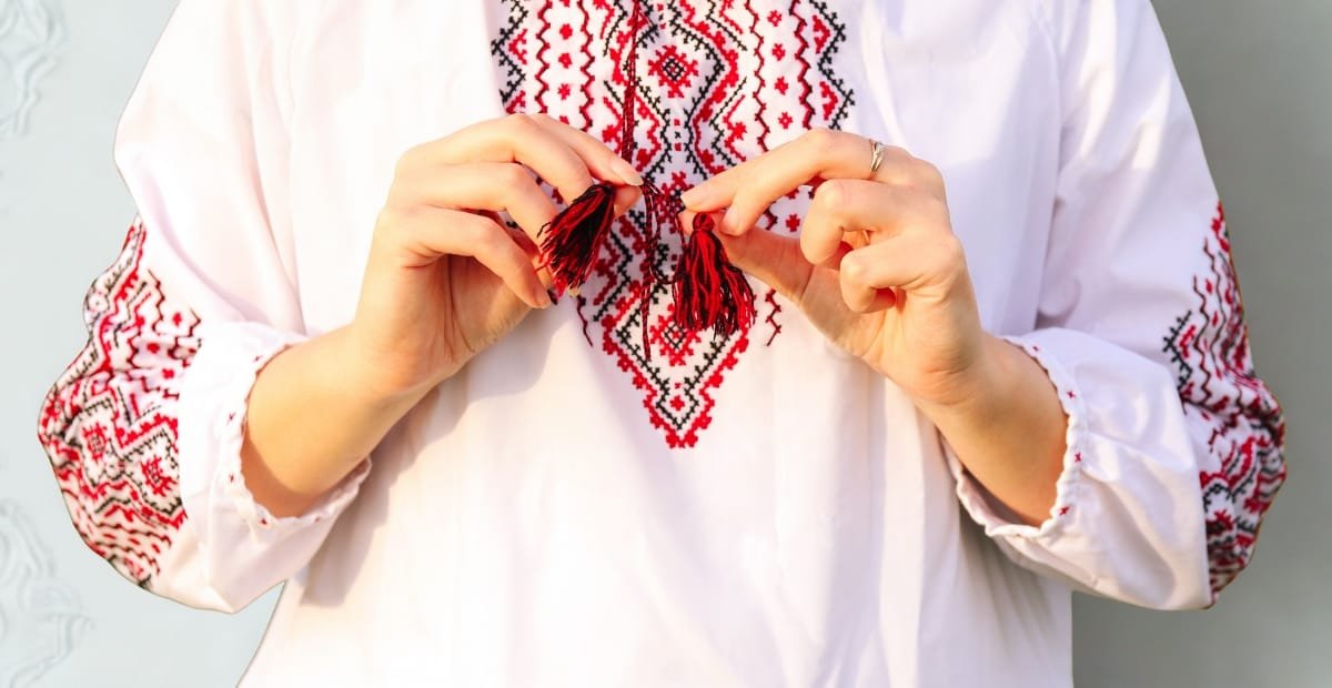 A person wearing a white embroidered blouse, holding a small red and black embroidered tassel with both hands.