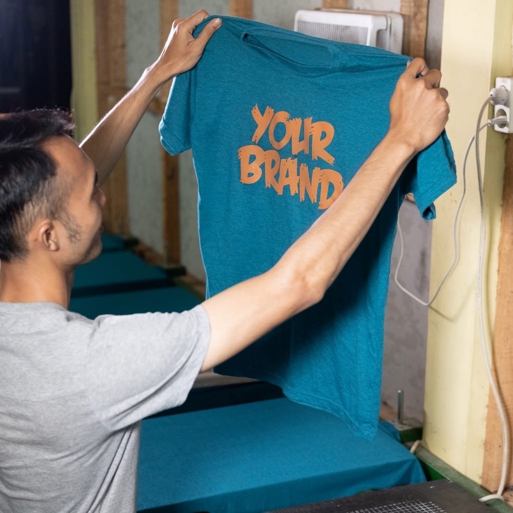 Man holding a blue t-shirt with bold orange text reading "YOUR BRAND".