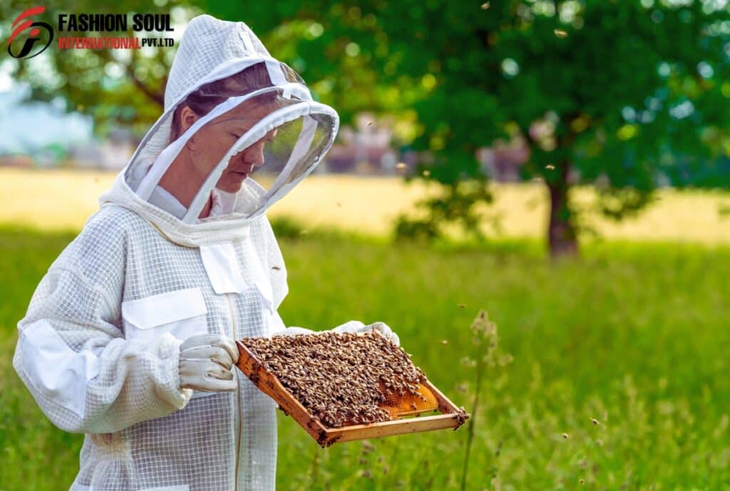 A beekeeper in protective white gear holds a frame full of bees outdoors, with a green field and trees in the background.