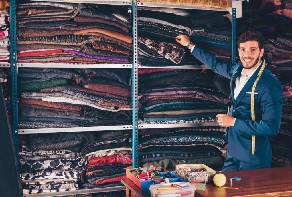 A cheerful man in a blue suit with a measuring tape around his neck is organizing fabric rolls on shelves in a textile shop. He is smiling and reaching for a piece of fabric, with sewing supplies on the table in front of him.