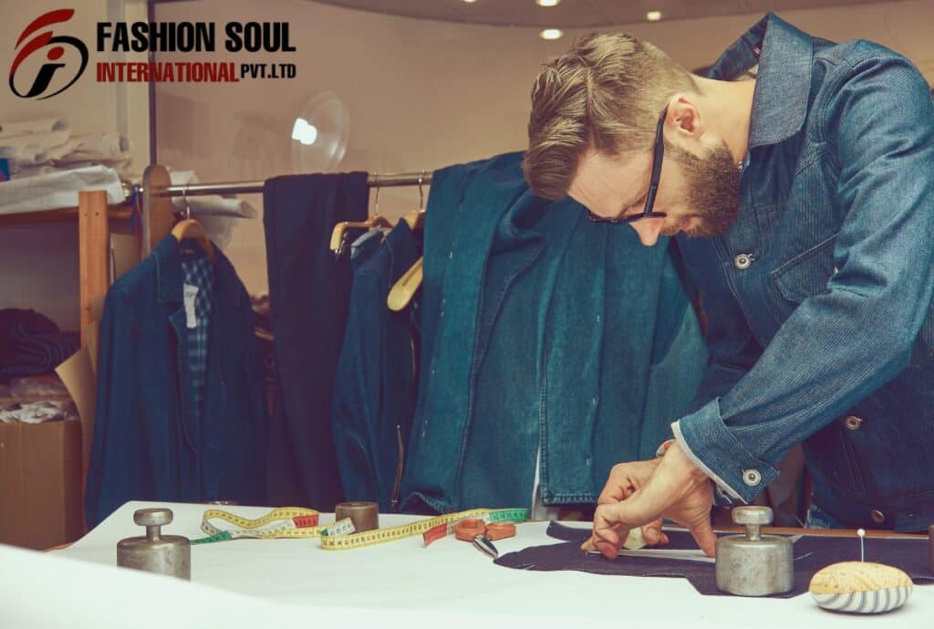 A man with glasses and a beard is leaning over a sewing table, working on a piece of black fabric. Behind him, there is a clothing rack with several denim jackets. The workspace has fabric measuring tapes, weights, and pins on the table, with shelves of folded clothes in the background. The logo in the top left corner reads "FASHION SOUL INTERNATIONAL PVT. LTD."