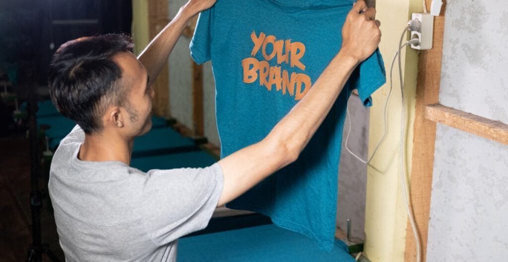 A man in a light gray T-shirt is holding up a blue T-shirt with orange text that reads “YOUR BRAND” as he threads it onto a display hanger or mannequin. He is indoors, near a wall with exposed studs and a power outlet.