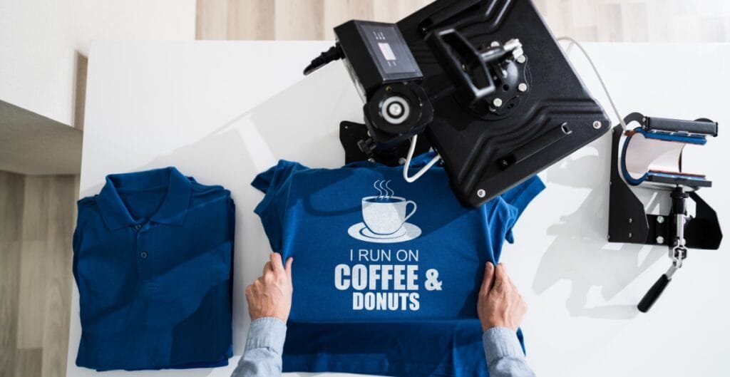 A person is pressing a blue T-shirt with the text "I RUN ON COFFEE & DONUTS" at a printing press, with folded blue shirts nearby on a white table.