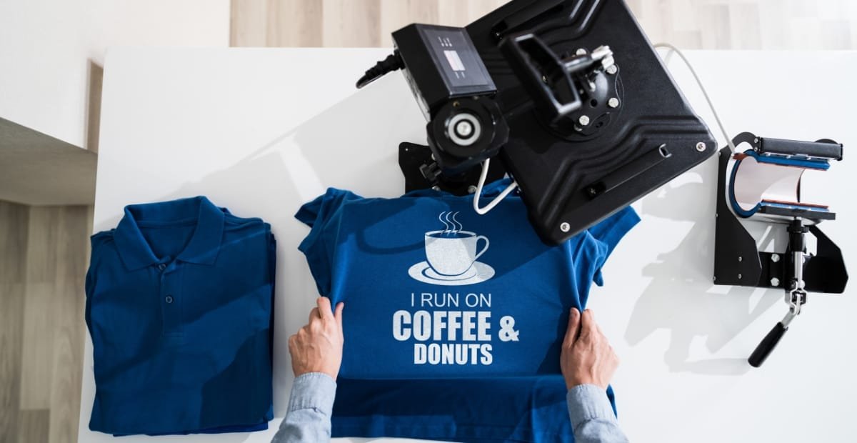 A person is pressing a blue T-shirt with the text "I RUN ON COFFEE & DONUTS" at a printing press, with folded blue shirts nearby on a white table.