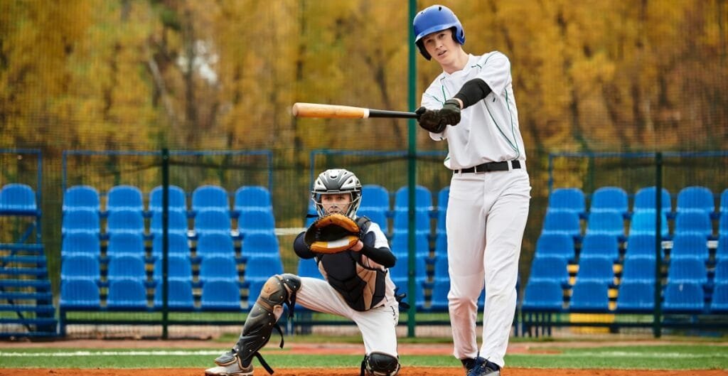 A young baseball batter in a white uniform and blue helmet swings at a pitch, while the catcher crouches behind him in gear, on a field with blue seats and autumn-coloured trees in the background.