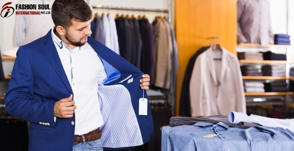 A man trying on a blue blazer inside a clothing store. He is holding the blazer open to show the lining on the inside, which has a light blue and white striped pattern. The store has racks of clothing in the background, and there are folded clothes on the table in front of him.