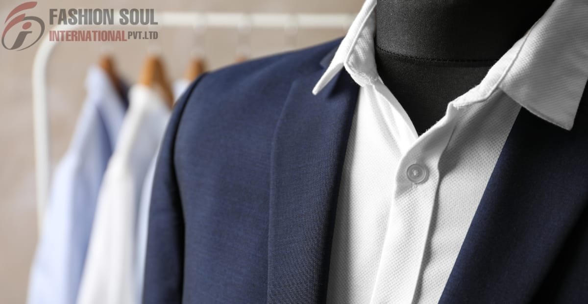A close-up view of a dark blue suit jacket worn over a white dress shirt on a mannequin, with blurred shirts hanging on a rack in the background. A logo reading “FASHION SOUL INTERNATIONAL PVT. LTD” appears in the top-left corner.