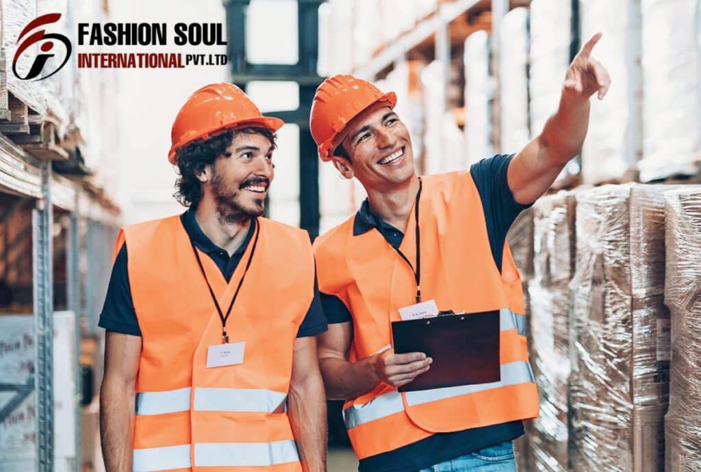 Two warehouse workers wearing orange safety vests and helmets are smiling and discussing inventory. One of them is holding a clipboard, and they are surrounded by stacked pallets in a well-lit warehouse.