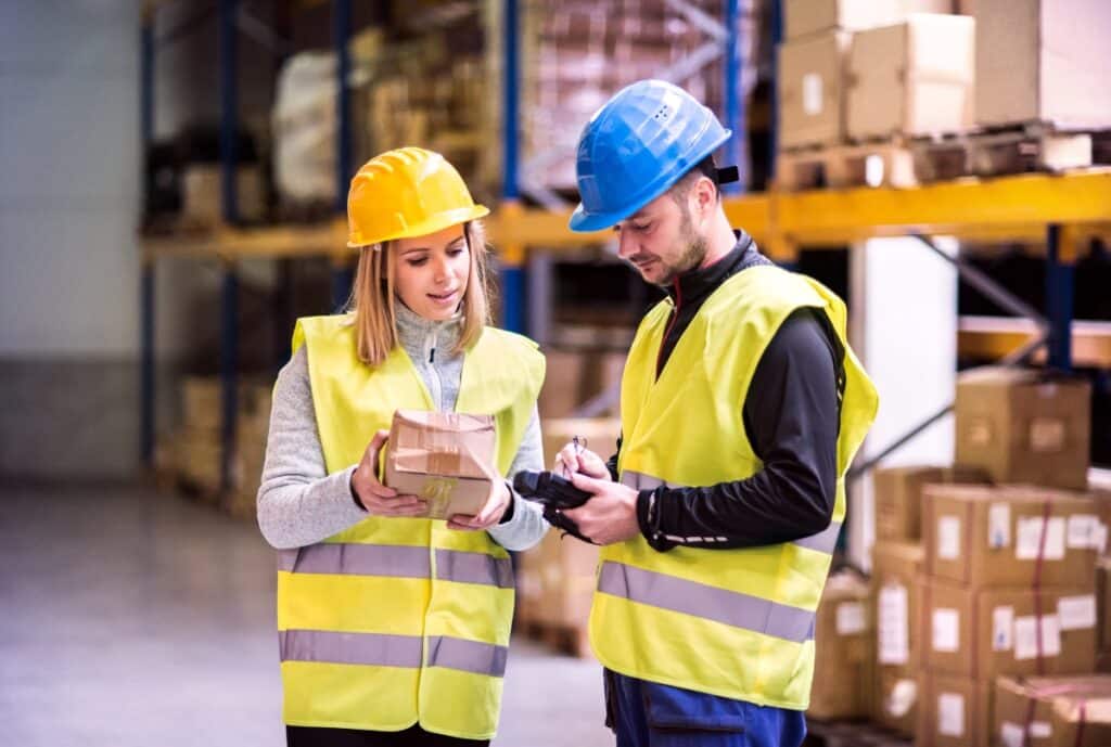 Two warehouse workers, a woman wearing a yellow safety helmet and vest and a man wearing a blue safety helmet and vest, are examining a package together in a storage area filled with shelves of boxes.