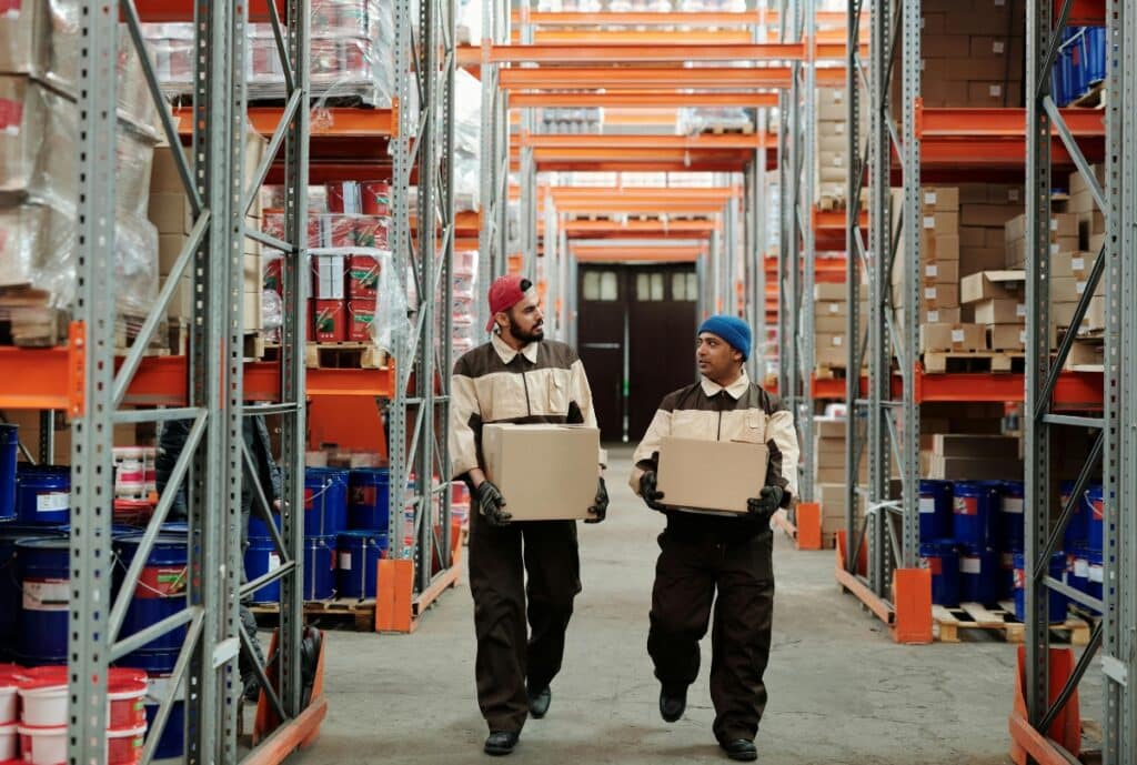 Two male warehouse workers, wearing protective gloves and uniforms with colored caps, are walking down an aisle carrying cardboard boxes in a large industrial storage area.