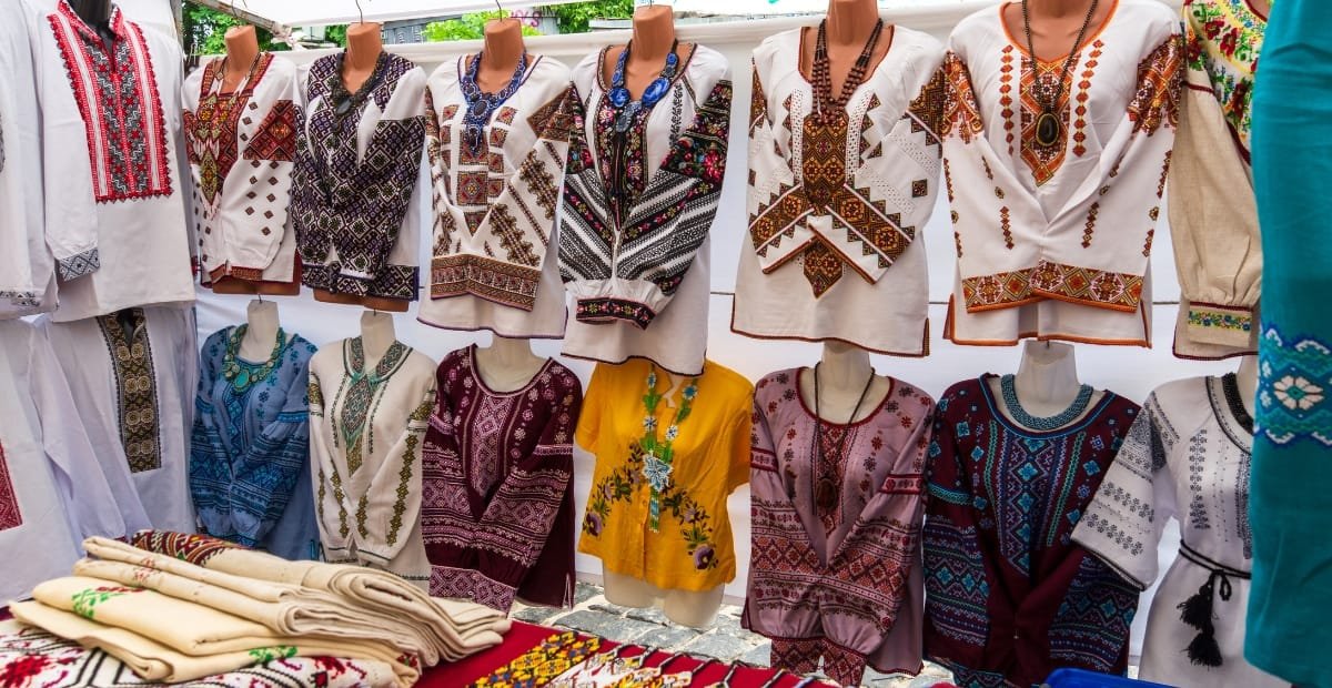 A market stall displaying a variety of traditional embroidered tunics and blouses on bust mannequins, with folded fabrics laid out on a table in front.