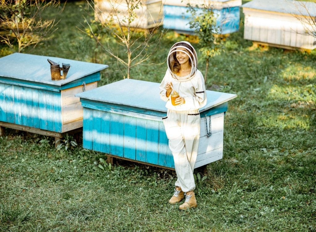 A woman dressed in a stylish beekeeping suit with a modern, fashionable touch, holding a jar of honey outdoors near blue and white bee hives.