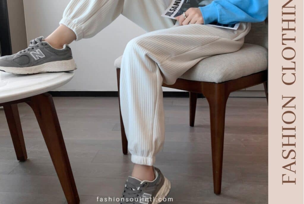 A person is seated on a wooden chair with one leg extended and resting on a small table. They are wearing light-colored, striped loose pants, gray New Balance sneakers, and a bright blue top. The scene appears to be indoors with a wooden floor and minimal background.