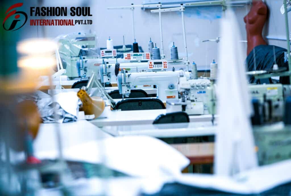 A sewing workshop with multiple industrial sewing machines on a long worktable, some fabric and mannequin parts visible. The workspace appears organized and ready for garment production. The logo "FASHION SOUL INTERNATIONAL PVT. LTD" is displayed in the top left corner.