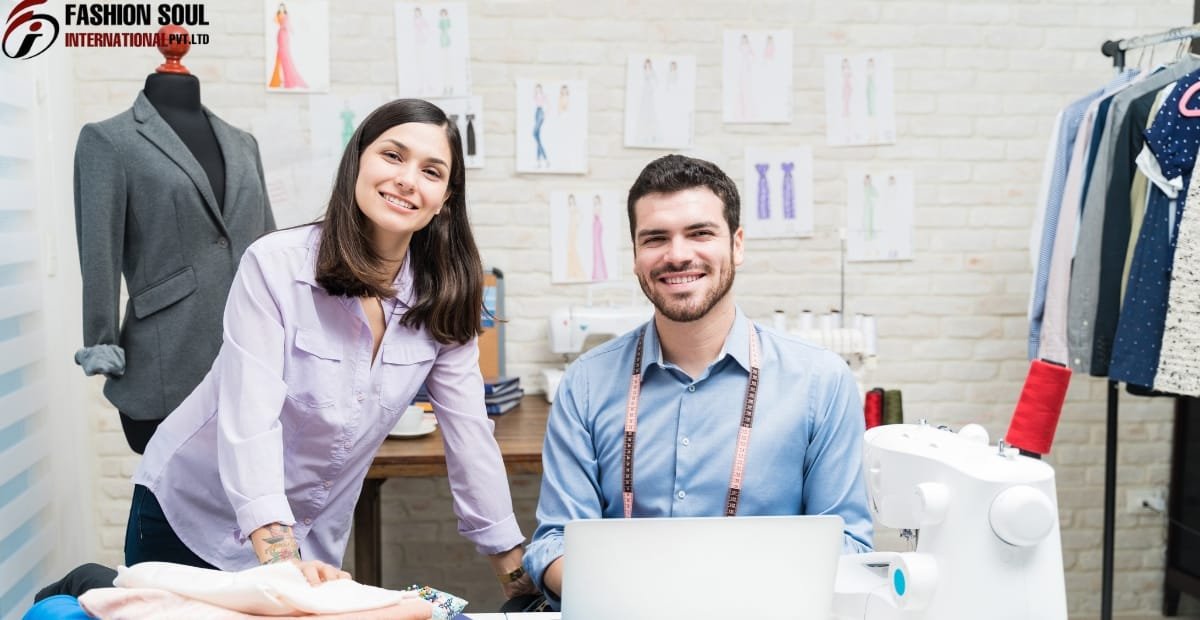 A woman and a man smiling at the camera inside a fashion design studio, with sewing equipment, fabric spools, and fashion sketches on the wall behind them.