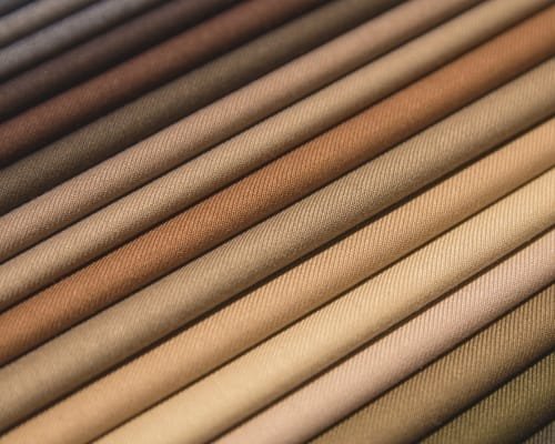 A close-up view of multiple rolled fabrics in a diagonal arrangement, showcasing a neutral color palette from dark browns to light beiges. The fabrics have visible woven textures and subtle shading, creating a gradient of earthy tones.
