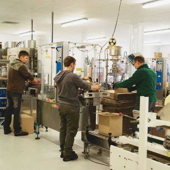 A modern factory workspace with three workers standing along a glass-enclosed packaging line. They are handling products on a metal conveyor belt, with cardboard boxes and packaging materials nearby. Industrial equipment, control panels, and overhead lighting fill the bright, clean facility.