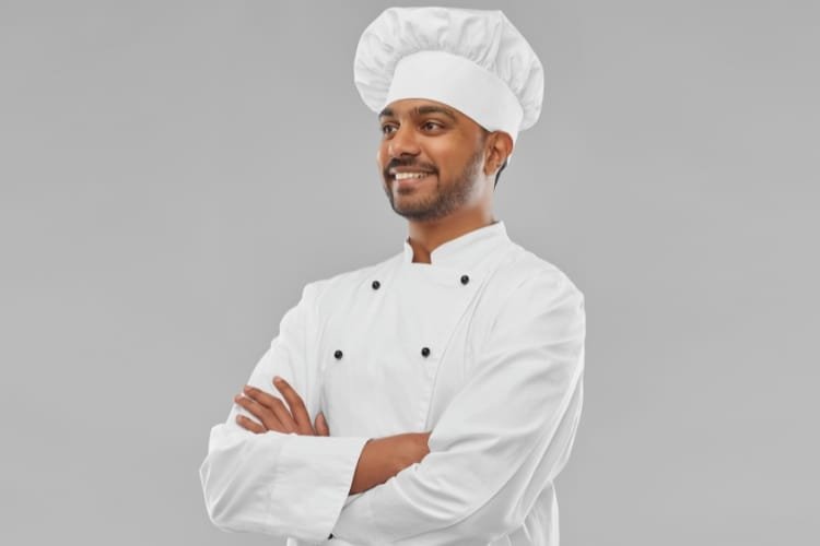  A cheerful male chef wearing a white chef’s hat and jacket, standing with arms crossed against a light gray background.