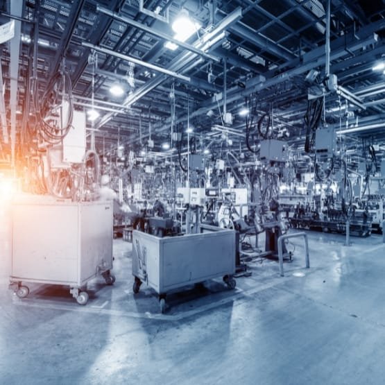 A spacious industrial factory floor filled with machinery, workstations, and overhead equipment. The scene shows a row of carts and various manufacturing stations, with bright lighting and a cool blue tone. A warm glow to the left suggests sunlight or illumination. The environment appears organized and busy, typical of a large-scale production facility.