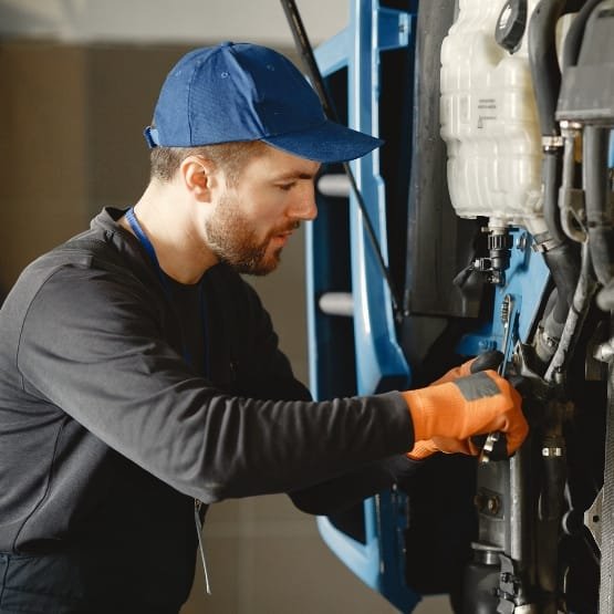 A male maintenance technician wearing a blue cap and orange gloves is repairing or assembling machinery. He is focused on a component, using a tool, with a large piece of industrial equipment and a white reservoir in the background.